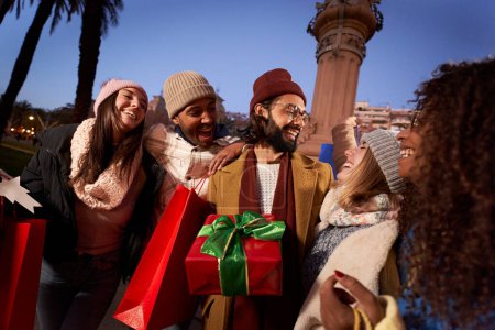 Photo for Excited smiling group multi-ethnic people holding colorful shopping bags gifts. Laughing young millennial friends enjoying winter night outdoor together community. Christmas vacation sale consumerism - Royalty Free Image