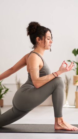 Photo for Vertical view of young caucasian girl in grey sportswear doing yoga. One-legged posture King Pigeon pose or Eka Pada Rajakapotasana. Calm woman controlling breath, meditating with eyes closed at home. - Royalty Free Image
