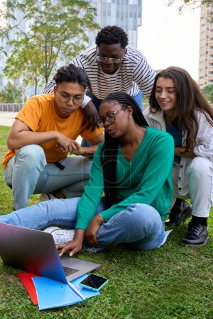 Foto de Vertical. Grupo de jóvenes estudiantes universitarios internacionales sentados en el césped fuera del edificio de la facultad. Amigos reunidos estudiando juntos con portátil en el campus - Imagen libre de derechos
