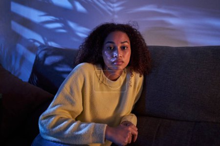 Photo for Portrait young African American woman looking serious on camera sitting alone on sofa at night home. Afro female posing with sad expression indoors. Photo of lonely, worried and dissociated black girl - Royalty Free Image