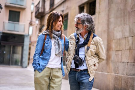 Photo for Cheerful senior attractive couple look at each other smiling on tourist street. Retirees enjoying leisure free time together on vacation outdoors. Mature marital relationship and pensioners holiday - Royalty Free Image