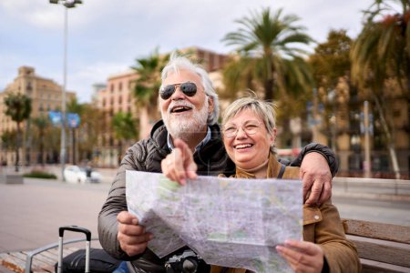 Senior pareja de turistas caucásicos sonriendo con mapa de viaje en las manos mirando y señalando lugares de interés. Ancianos marido y mujer sentados en el banco en la calle de la ciudad disfrutando de vacaciones. Turismo de adultos 
