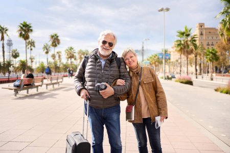 Photo for Smiling Caucasian mature tourist couple standing posing looking at the camera in the street with their luggage. Husband and wife enjoying their retirees holidays wearing winter clothes on sunny day - Royalty Free Image