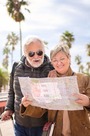 Photo for Vertical elderly Caucasian tourist couple smiling holding travel map looking together interest places. Senior husband and wife standing in city enjoying pensioner vacation. Adult tourism people - Royalty Free Image