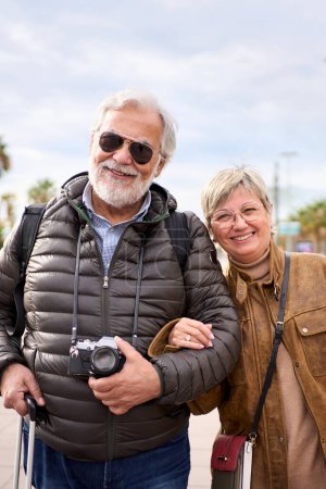 Photo for Vertical. Smiling Caucasian mature tourist couple standing posing looking at camera in the street with their luggage. Husband and wife enjoying their retirees holidays wearing winter clothes - Royalty Free Image