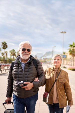 Photo for Vertical smiling Caucasian mature tourist couple standing posing looking at camera in street with their luggage. Husband and wife enjoying their retirees holidays wearing winter clothes on sunny day - Royalty Free Image