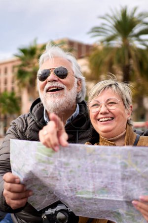 Photo for Vertical senior Caucasian tourist couple smiling holding travel map looking and pointing interest places. Elderly husband and wife sitting on bench in city enjoying vacation. Adult tourism people - Royalty Free Image