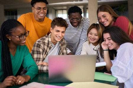 Fröhliche junge Gruppe von Studenten mit Laptop in der Cafeteria auf dem Campus mit seinen Kommilitonen, die an einem Projekt arbeiten. Lächelnde multiethnische Menschen, die gemeinsam auf dem Fakultätsgebäude lernen