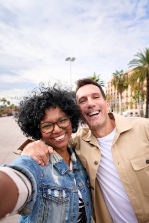 Vertikal reifes Touristenpaar macht Selfie und umarmt sich. Mann und schwarze Frau lächeln verliebt in die Kamera und genießen ihre Reisen in der europäischen Stadtstraße. Erwachsenenbesichtigung 