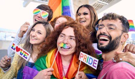 Group cheerful young friends taking fun selfie for social media celebrating gay pride day outdoors. Diverse people wearing colorful props and rainbow flags. Concept of LGBT community and generation z.