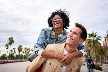 Reife multirassische freudige Touristenpaar huckepack stehend im Freien. Fröhliche schwarze Frau und kaukasischer Mann lächelnd, die auf der Straße der Stadt spazieren gehen und fröhliche Ferien genießen. Kopierraum, Hintergrund Palmen