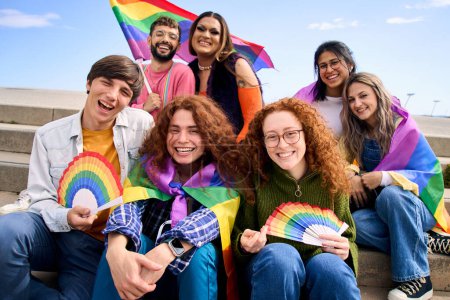 Portrait group diverse people smiling for gay pride day photo. Young happy friends gathered celebrating with rainbow flags LGBT party sitting outdoor. Generation z and positive equal relationships.