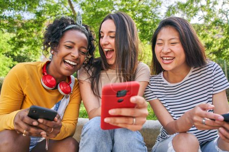 Téléchargez les photos : Trois joyeuses femmes multiraciales utilisant le téléphone assis à l'extérieur en été. Excité fille caucasienne montre quelque chose de drôle sur mobile à de bons amis. Jeune femme riant de surprise en regardant le dispositif d'écran - en image libre de droit