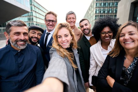 Selfie eines gut gelaunten Teams diverser Geschäftsleute im formellen Anzug, die lächelnd in die Kamera schauen, die sich vor dem Werksgebäude versammelt haben. Positive Emotion Teamarbeit feiert gemeinsam Erfolg