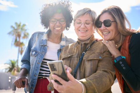 Foto de Grupo de mujeres multirraciales de mediana edad que sonríen mirando y usando el smartphone en la mano en la calle de una ciudad, buscando lugares y disfrutando de sus vacaciones en un día soleado de otoño - Imagen libre de derechos