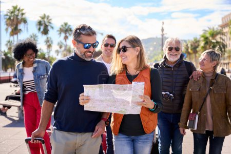 Couple of middle-aged walking and looking and holding a travel map with a group diverse tourist friends with luggage. Tourism of people enjoying their holidays on a European city street