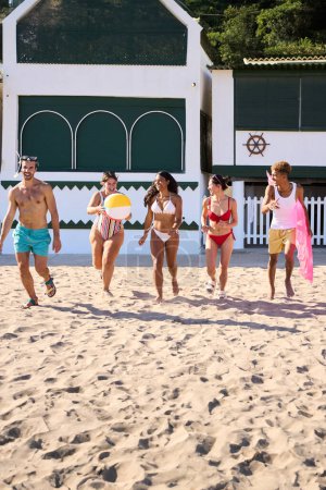 Vertikales Foto einer Gruppe aufgeregter Freunde, die in Badebekleidung am Strand entlang laufen. Junge multiethnische Menschen haben Spaß im Sommerurlaub am Strand
