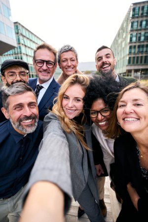 Vertical selfie of a cheerful team of diverse business people in formal suit looking smiling at camera gathered outside the work building. Positive emotion team work celebrating success together