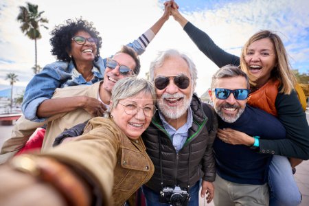 Group diverse funny mature tourist friends posing piggyback with hands joined in air taking photo selfie together with front camera on travel outdoor. Adult happy six excited people enjoy holidays 
