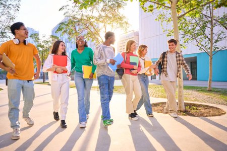 Foto de Estudiantes internacionales felices caminando por el campus universitario. Jóvenes amigos multiculturales sonriendo y mirándose con mochilas y cuadernos al aire libre - Imagen libre de derechos