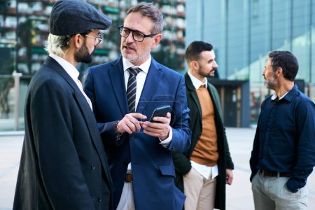 Group of Caucasian business men gathered talking serious about issues using and holding mobile phone on break work outdoor. Stylish professional males together conversing quiet outside office building