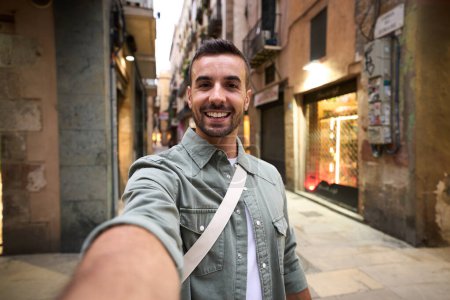 Photo for Young adult happy Caucasian man taking smiling selfie on Madrid touristic city street in background. Millennial handsome tourist takes lighthearted photo outdoors on his vacation trip in Europe - Royalty Free Image