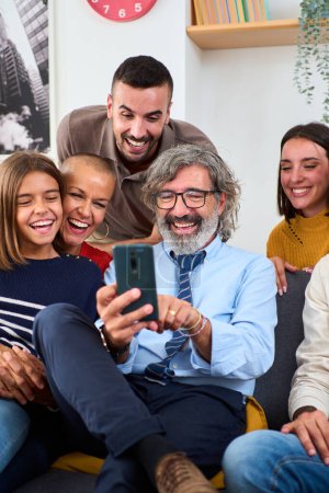 Vertikal. Kaukasische Mehrgenerationenfamilie versammelte sich im Wohnzimmer auf dem Sofa sitzend mit dem Handy. Glückliche Menschen lächeln mit lustigem Gesichtsausdruck und genießen und gucken gemeinsam Videos in der Zelle zu Hause