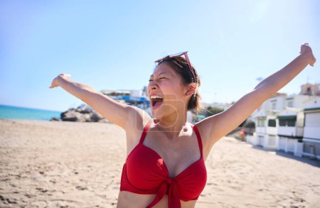 Porträt einer jungen asiatischen Chinesin im roten Bikini mit breitem Lächeln, während sie die Arme am Strand hebt. Glückliches und fröhliches Mädchen in den Sommerferien. Reisen der Generation Z