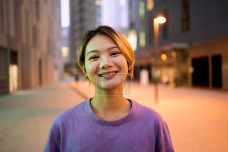 Portrait schöne asiatische schöne junge Frau lächelt mit freundlichem Gesichtsausdruck und schaut freudig in die Kamera draußen. Generation Z hübsche Chinesin mit blonden Haaren posiert in der Abenddämmerung fröhlich in der Innenstadt 