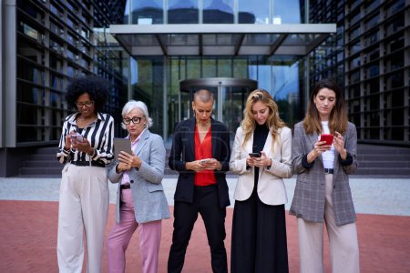 Group of diverse ages and races of business women in formal wear walking using mobile phone. Technology addicted serious adult professional females outside the office building with cells in their hand