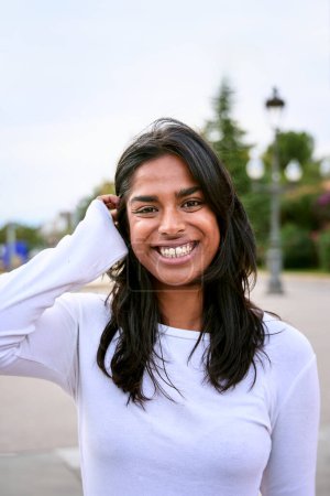 Vertical Portrait of beautiful Young Indian Woman Looking at Camera and Smiling outdoors city street. Happy adult girl of India. Female person with positive facial expression beautiful skin tilts head