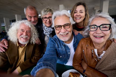 Feliz hombre de pelo gris maduro tomando selfie de seniors grupo amigos alegres caucásicos posando juntos sentados en la cafetería. Personas mayores mirando sonriendo a la cámara con bocadillos en la mesa hogar de ancianos 