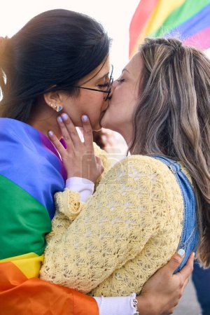 Vertical. Two young loving girls kissing on gay pride festival day outdoors. Group of friends celebrating lgbt party on background with rainbow flag. Generation z and types of female sexuality.