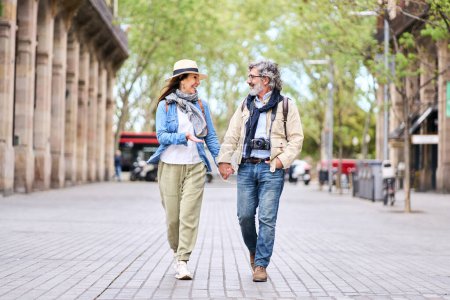 Happy adult Caucasian tourist couple looking at each other walking holding hand for street in city. Mature people in love enjoying romantic getaway. Positive relationships and spring retiree holidays