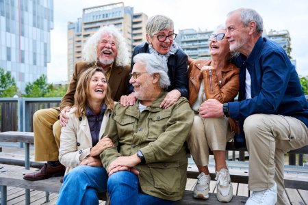Eine Gruppe älterer Kaukasier, die zusammen auf einer Bank in der Stadt sitzen und lachen. Glückliche alte Freunde mit grauen Haaren versammelten sich und hatten Spaß im Freien. Lebensstil gut gelaunter älterer Menschen genießt sonnigen Herbsttag