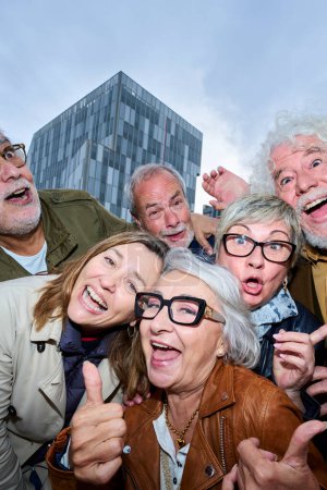 Vertikal. Lustiges Porträt einer Seniorengruppe kaukasischer Menschen, die zusammen im Freien gestikulierend posieren. Ältere Freunde blicken freudig in die Abenddämmerung der Stadt. Lebensstil glücklich reift Partner