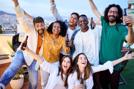 Group of diverse excited friends celebrating with beers at barbecue party in attic. Portrait of young people posing raising arms on terrace in evening in open air. Cheerful colleagues enjoying time.