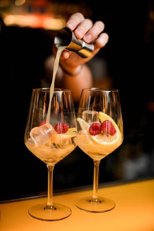 Photo for Two transparent glasses with ice cubes and lemon slices and raspberry on bar counter and hand of bartender pours drink from steel jigger into glass - Royalty Free Image