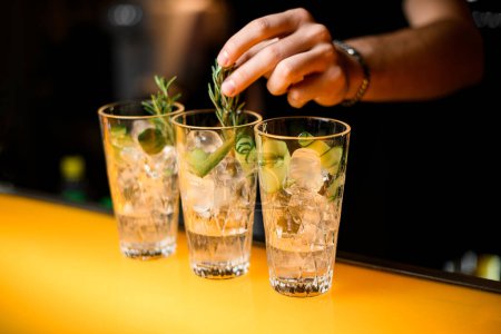 Foto de Enfoque selectivo en vasos con cóctel gin tonic que mano de camarero masculino precisa decorar con rama de romero - Imagen libre de derechos