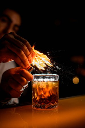 Foto de Increíble vista de cristal anticuado con bebida alcohólica en el mostrador de la barra y mano masculina asperja por la ralladura y lo pone en el fuego - Imagen libre de derechos