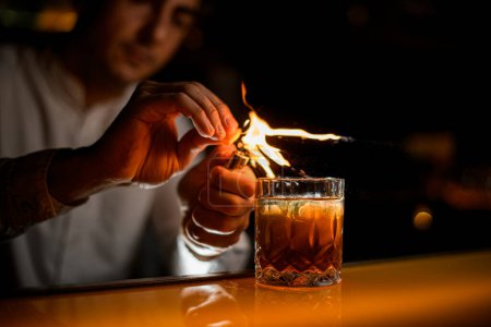 Foto de Gran vista de cristal anticuado con bebida alcohólica en el mostrador de la barra y mano masculina asperja por la ralladura y lo pone en el fuego - Imagen libre de derechos