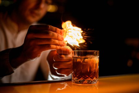 Foto de Vista de cristal anticuado con bebida alcohólica en el mostrador de la barra y mano masculina asperja por la ralladura y lo pone en el fuego - Imagen libre de derechos