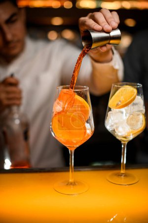 Photo for Glass with ice cubes and orange slices on the bar counter and hand of the bartender pours drink from steel jigger into glass - Royalty Free Image