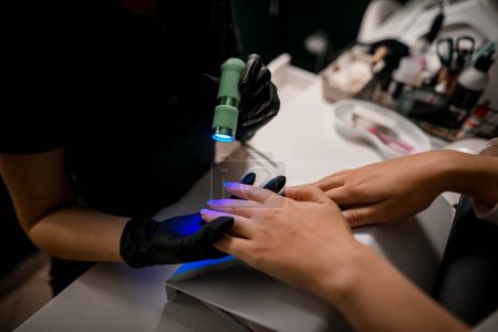 Téléchargez les photos : Vue rapprochée des mains féminines de manucure qui utilisent une lampe ultraviolette ou UV pour durcir le couvercle supérieur du vernis à ongles. - en image libre de droit