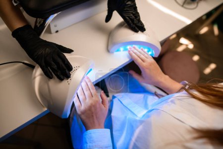 Téléchargez les photos : Les mains féminines utilisent la lampe ultraviolette ou UV dans le salon des ongles pour sécher les ongles. Gel poli processus de manucure - en image libre de droit