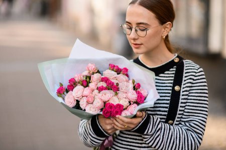 Photo for Beautiful woman holds bouquet of bright pink roses in wrapping paper in her hands and look at it - Royalty Free Image