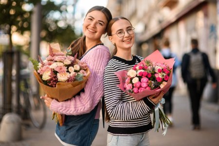 two happy women with lush flower bouquets with fresh roses in wrapping paper in their hands outdoors