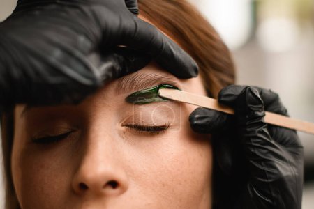 Primer plano del proceso de aplicación de cera verde a la piel en las cejas femeninas para eliminar el cabello no deseado. Corrección profesional en salón de belleza.