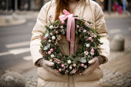 großer runder Weihnachtskranz aus Tannenzweigen, dekoriert mit rosa Glitzerkugeln und Pflanzen und Verbeugung in weiblichen Händen