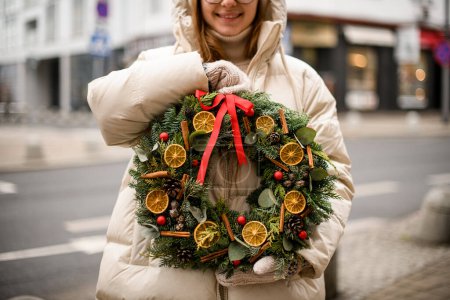 Téléchargez les photos : Belle couronne de Noël ronde décorée de tranches d'orange sèches boules de cannelle et ruban dans les mains féminines - en image libre de droit